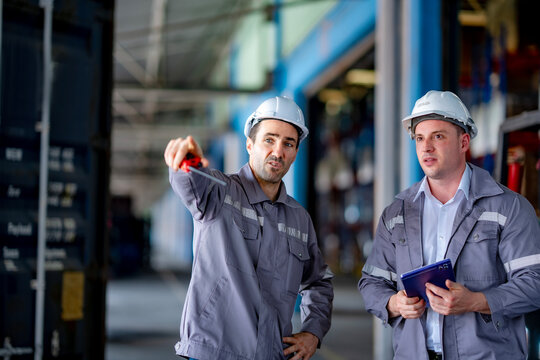 Two factory workers discuss shipment plans in front of a warehouse. A loading truck and storage shelves in the background show active supply chain operations. - Powered by Adobe