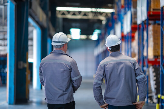 Two industrial workers in uniform and hard hats inspecting warehouse facility. Rear view of team walking through storage area for quality control, safety checks, and inventory assessment. - Powered by Adobe