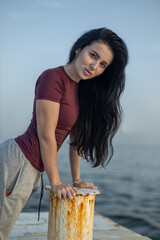 woman stands on a dock, leaning on a rusted pole while gazing at the water She wears a maroon top...
