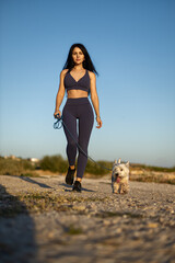 brunette woman strolls along a sunny beach with her white dog She engages in skin care, focusing on...