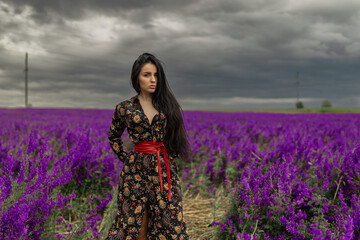 brunette woman wears a floral dress with a red belt while posing in a vibrant field of delphiniums...