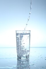 Pouring water into glass at mirror table against light blue gradient background, closeup