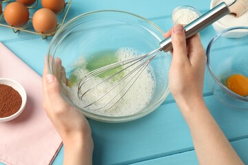 Woman whisking egg whites in bowl at light blue wooden table, closeup