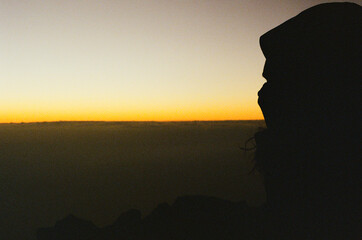 A silhouette of a person gazing at a colorful gradient sunset on a volcano above the clouds, evoking serenity and introspection. Capturing the transition between day and night on a tranquil horizon.
