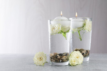 Burning candles in glasses with water, stones and rose flowers on light grey table, closeup. Space...