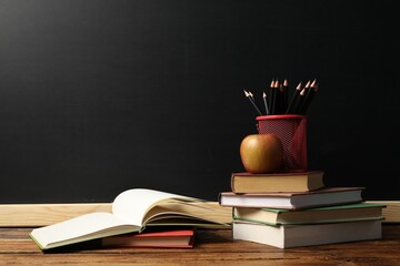 Different books, pencils and apple on wooden table near blackboard
