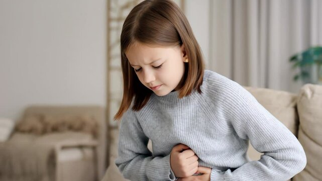 A young girl clutches her stomach in discomfort, sitting on a couch at home.