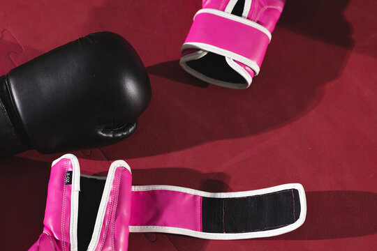 Black boxing glove lying on red padded ring floor with two pink gloves exposing unfastened strap