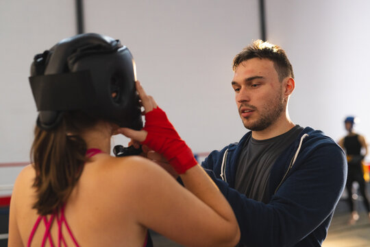 Male coach adjusting headgear for female trainee with red hand wraps at boxing gym, copy space