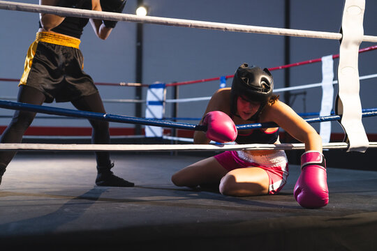 Two female boxers kneeling and standing inside boxing ring at gym with gloves headgear ropes