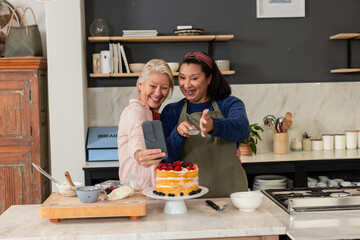 Diverse senior female friends taking smartphone selfie at kitchen counter near piping bag and cake