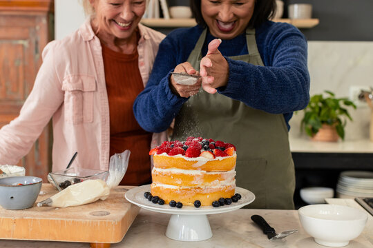 Diverse senior female friends sprinkling sugar over cake on stand at kitchen counter, copy space