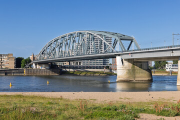 The majestic Nijmegen Railway Bridge, a vital steel arch bridge, spans the Waal River on a bright summer day, with the city's modern skyline visible in the background.
