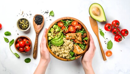 Hands holding a wooden bowl filled with a healthy vegan meal of quinoa, avocado, tomatoes, tofu, and greens, with other ingredients around.