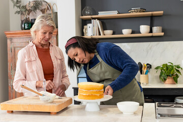 Diverse female friends decorating three-layer sponge cake on island in home kitchen with spatula