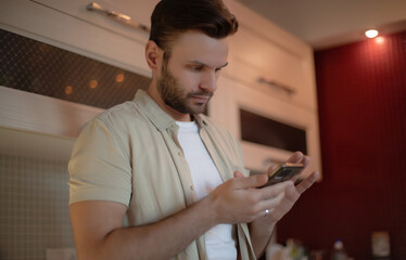 Young man in casual clothes stands in a kitchen, looking at his smartphone with a focused and...