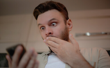 Shocked young man in casual clothes looks at his smartphone with wide eyes, covering his mouth with...