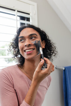 Fototapeta Non-binary person applying dark facial mask to cheek with makeup brush by window in bathroom