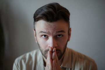 Close-up of a young man in casual clothes with hands pressed together near his face, looking...