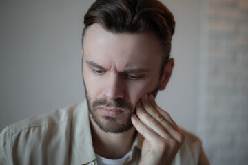 Young man in casual clothes indoors touching his cheek with a concerned, pained expression, showing...