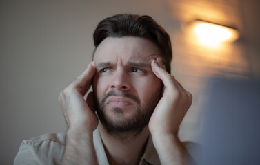 Close-up of a young man with beard and beige shirt holding his head with both hands, looking...