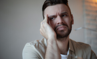 Young man in casual shirt looking stressed and tired, touching his face with concern, sitting at...