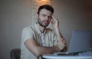 Frustrated man with eyes closed holding head in pain, sitting at desk with laptop, suffering from...