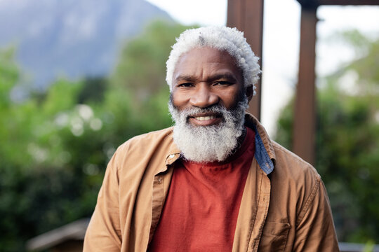 Senior African American man wearing red shirt tan jacket standing in yard with pergola and mountain