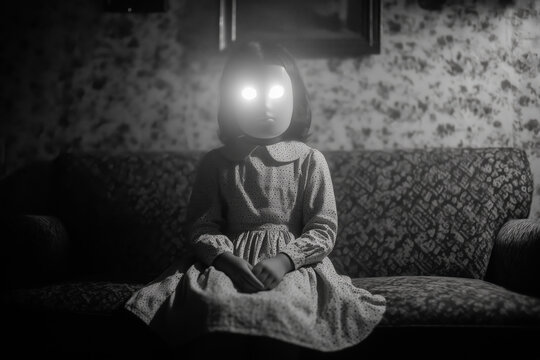 1960s Black and white film portrait of mysterious little girl in floral dress and illuminated blank papier-m&acirc;ch&eacute; mask, sitting on vintage flowery sofa in foggy room