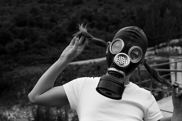 A young woman wearing a gas mask stands outdoors and playfully pulls her pigtails