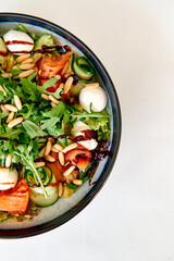 Fresh salad bowl with arugula, mozzarella, salmon, and pine nuts garnished with cucumbers on white background