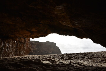 View from inside a cave at Praia do Amado, Algarve, Portugal. The sandy floor leads to waves crashing on the shore, framed by dramatic coastal cliffs.