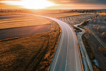 Fototapeta premium Aerial view of a winding road crossing colorful agricultural fields at sunset, creating a picturesque landscape