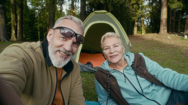 POV shot of mature Caucasian couple looking at camera recording video vlog while lying on grass near tent and sharing summer camping trip experience