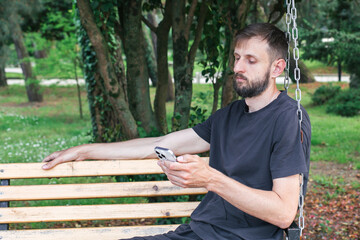 Fototapeta premium Man with beard sitting on wooden swing bench in park holding smartphone, looking at screen with blank expression, concept of digital addiction.