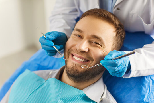 Dental health. Man sits comfortably in dentists chair, showing off white smile that exudes confidence. Dentist holds special tools in his hands for preventive and therapeutic examination of teeth.