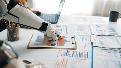 A robotic hand closely examines business charts and graphs on a clipboard in a modern office. The...