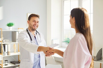 Obraz premium Smiling male doctor in white medical uniform meets to consult female patient, hand shaking, male GP welcomes in cabinet to talk, discuss checkup results with woman client at meeting in modern hospital