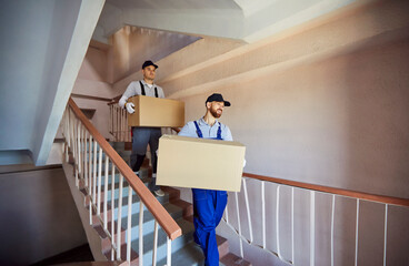 Men movers carrying big packed cardboard boxes with belongings, posing on stairs in entrance, help...