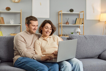 Fototapeta premium Family, relationship and technology concept. Couple enjoy spending free time together at home surf internet via laptop, watch movie or post photos in social networks sit on comfortable sofa