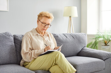 Focused alone busy senior woman sitting on sofa writing in copy book, fixing message, leading...