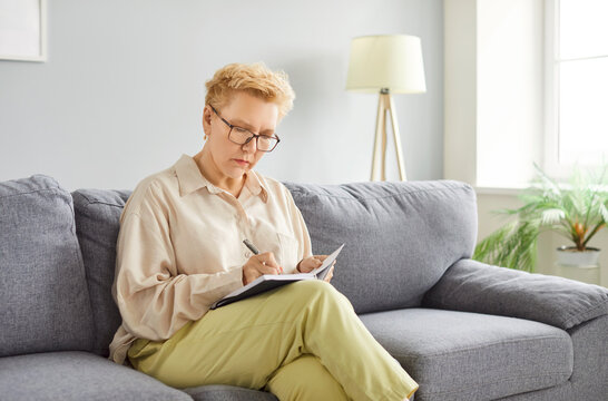 Focused alone busy senior woman sitting on sofa writing in copy book, fixing message, leading correspondence with relatives, searching information, planning new day, relaxing weekend time, thinking  - Powered by Adobe