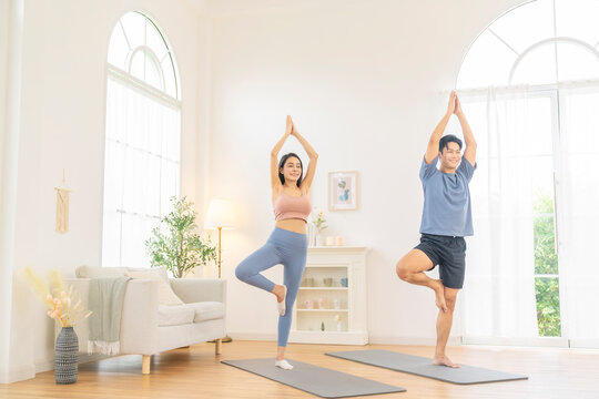 Happy Asian family couple practicing meditation yoga wellness exercise together in living room. Healthy people man and woman enjoy sport training workout body stretching yoga class on yoga mat at home