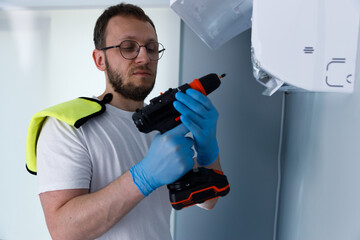 Fototapeta premium Man in Blue Gloves Holding a Power Drill While Preparing for Installation Tasks, Wearing a White T-Shirt with a Bright Yellow Towel on His Shoulder Indoors in a Modern Setting