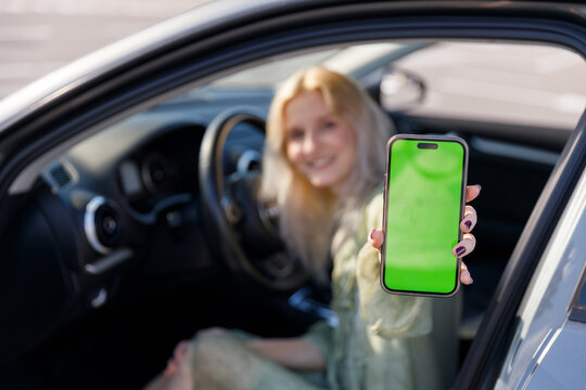 Young woman seated in a car, joyfully displaying her smartphone with a green screen, perfect for app developers and marketers seeking mockup visuals in an automobile setting.