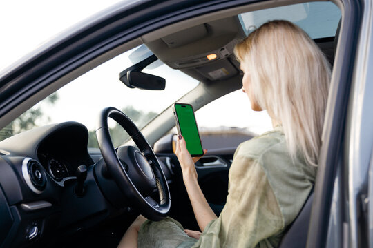 A Young Woman in a Car Holding a Smartphone with a Green Screen Background While Relaxing in the Driver's Seat on a Bright Day
