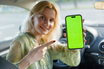 A Smiling Woman Inside a Car Holding a Smartphone with a Bright Green Screen, Engagingly Pointing...