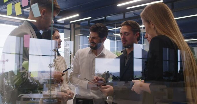 Young motivated team busy planning discussing creative startup ideas using sticky papers, employees looking at stickers, successful result, modern workers brainstorming behind modern glass office wall