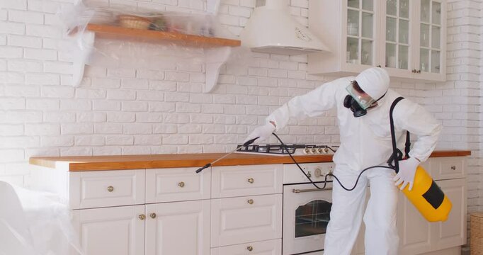 Man in protective suit and mask disinfecting kitchen during pandemic to kill bacteria. Termite pest control worker spraying and cleaning home from insects. Sanitary treatment concept