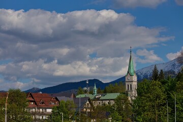 Church with green spire stands amid red-roofed homes and forest; snow-capped peaks frame tranquil village under cloudy skies.